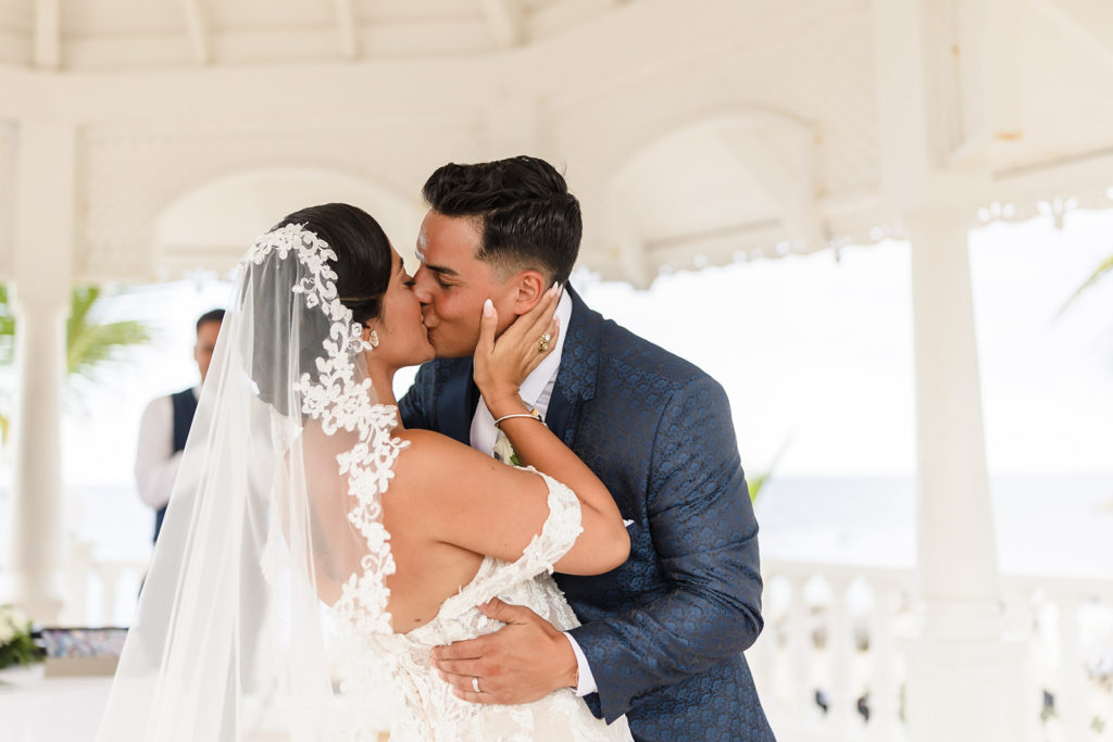 A bride and groom kiss at their Caribbean Destination Wedding photographed by TKM Photography located in Roatan, Honduras and Durham Region, Ontario, while her sister plays traditional wedding songs behind them. 