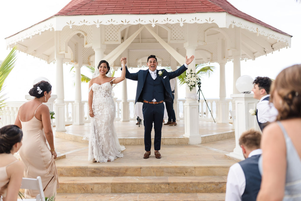 The bride and groom smile with their hands in the air after the ceremony at their Caribbean Destination Wedding photographed by TKM Photography located in Roatan, Honduras and Durham Region, Ontario, while her sister plays traditional wedding songs behind them. 
