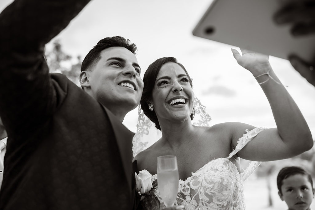 The bride and groom smile and wave at their virtual guests on a laptop at their Caribbean Destination Wedding photographed by TKM Photography located in Roatan, Honduras and Durham Region, Ontario, while her sister plays traditional wedding songs behind them. 