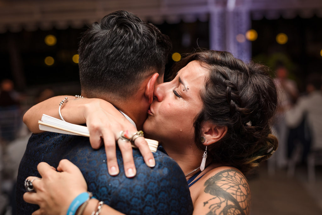 The groom's sister kisses his cheek while a tear runs down her cheek at a Caribbean Destination Wedding photographed by TKM Photography located in Roatan, Honduras and Durham Region, Ontario, while her sister plays traditional wedding songs behind them. 