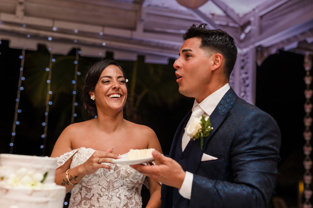A bride laughs after putting wedding cake on her husband's face at their Caribbean Destination Wedding photographed by TKM Photography located in Roatan, Honduras and Durham Region, Ontario, while her sister plays traditional wedding songs behind them. 