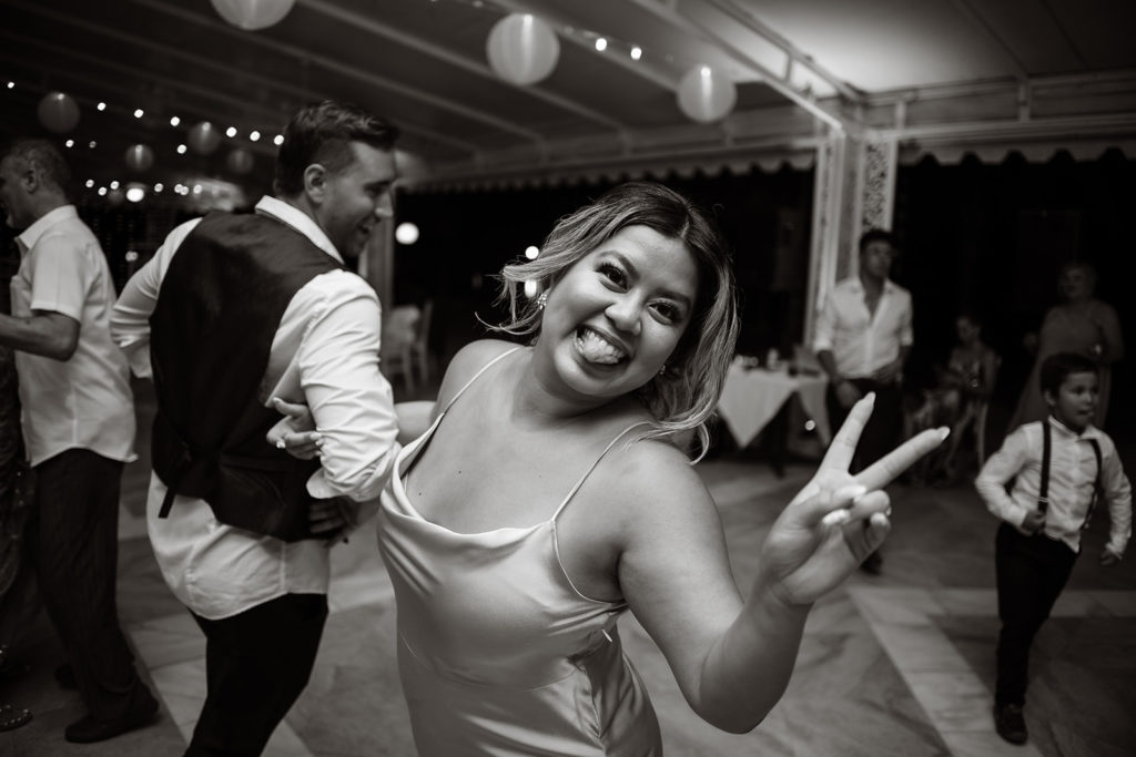 A bridesmaid dances in a circle with her husband like giving a peace sign and sticking her tongue out at a Caribbean Destination Wedding photographed by TKM Photography located in Roatan, Honduras and Durham Region, Ontario, while her sister plays traditional wedding songs behind them. 