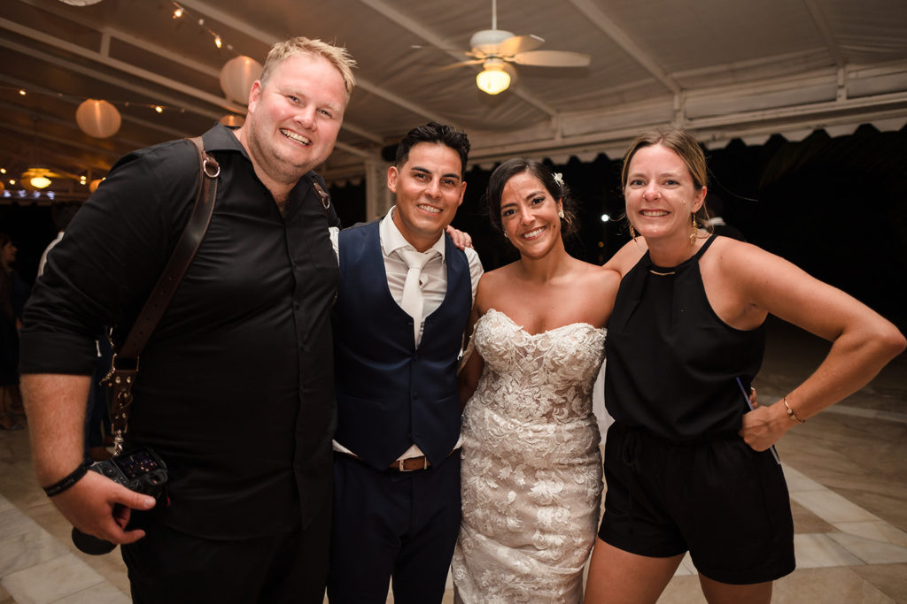 Kat and Taylor, wedding photographers of TKM Photography, smile with the bride and groom at their Caribbean Destination Wedding photographed by TKM Photography located in Roatan, Honduras and Durham Region, Ontario, while her sister plays traditional wedding songs behind them. 