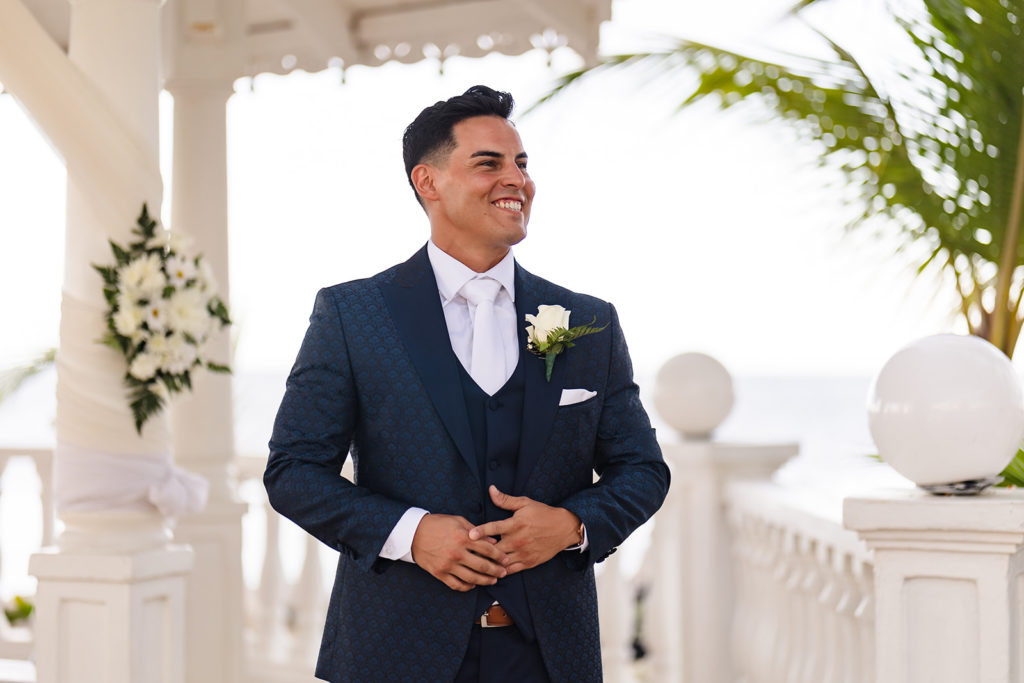 A groom smiles wide as he watches his bride walk down the aisle at a Caribbean Destination Wedding photographed by TKM Photography located in Roatan, Honduras and Durham Region, Ontario, while her sister plays traditional wedding songs behind them. 